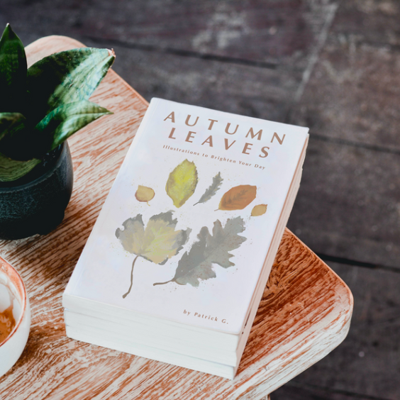 A photo of a small book on a wooden table. Text above reads 'Autumn Leaves' and image of seven leaves is digitally painted underneath.